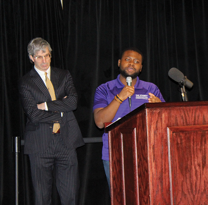 Man with purple shirt talking through a mic behind a stage podium with a man in a dark grey suit standing next to him.